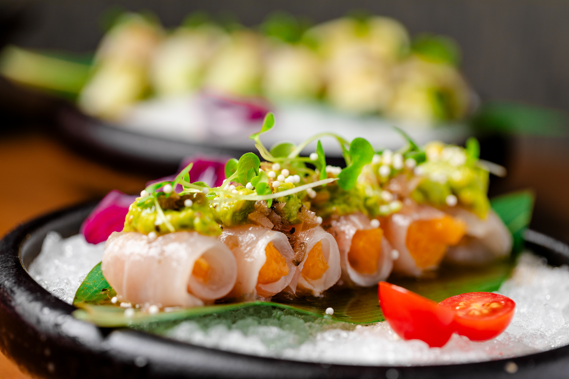 Close-up of yellowtail crudo rolls topped with wasabi avocado, microgreens and sesame over tomato and crushed ice