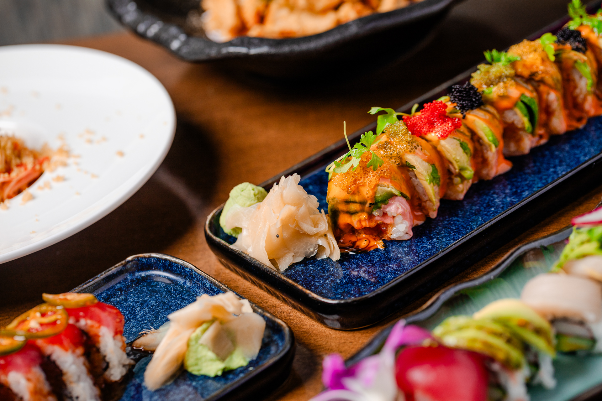 A long avocado-wrapped sushi roll drizzled with spicy sauce and topped with golden, red and black roe on a cobalt blue platter, with pickled ginger and wasabi alongside and other sushi plates nearby