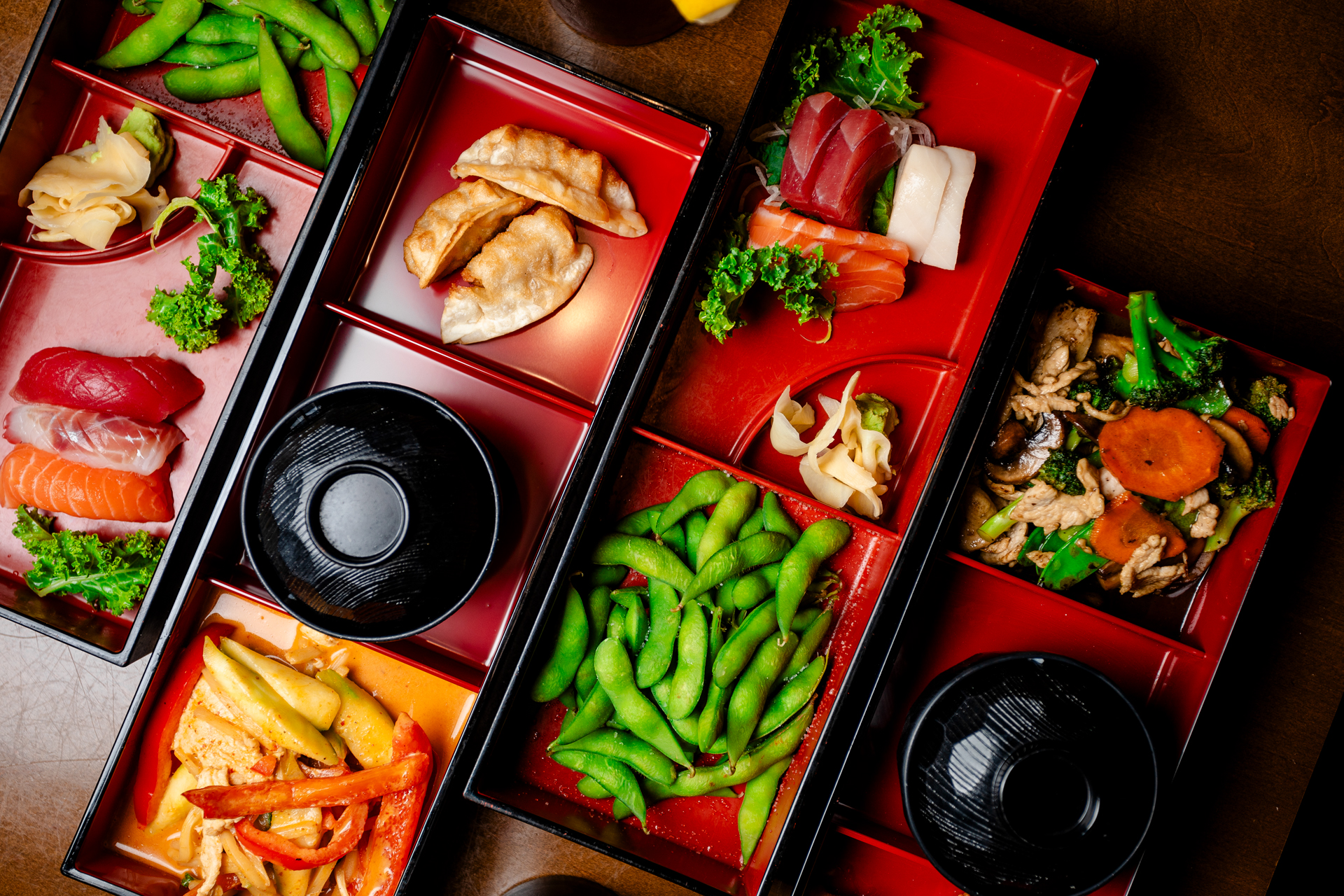 Red and black bento boxes with sushi, sashimi, edamame, dumplings, noodles and soup